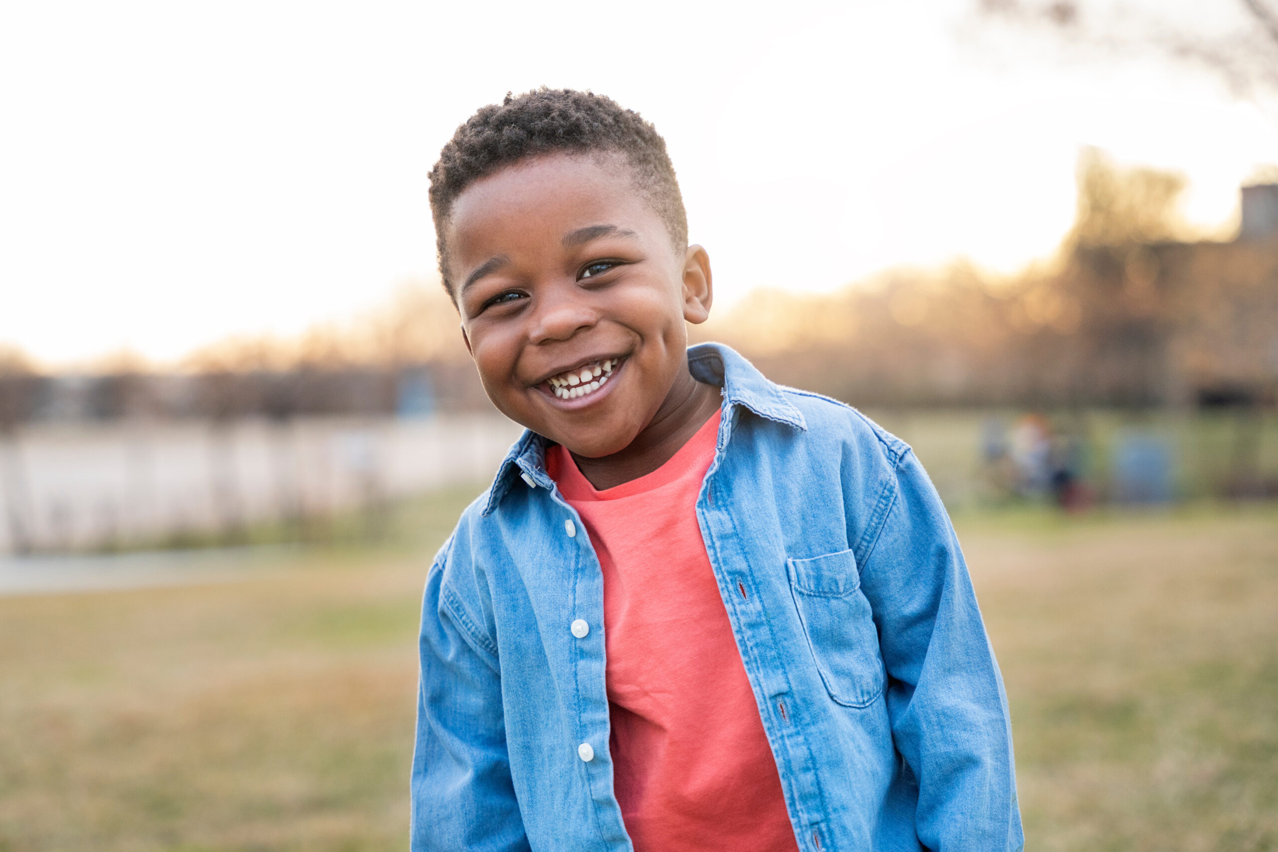 A young boy with short curly hair smiles brightly outdoors, wearing a light blue denim shirt over a coral t-shirt. The background shows a grassy area and soft sunlight.