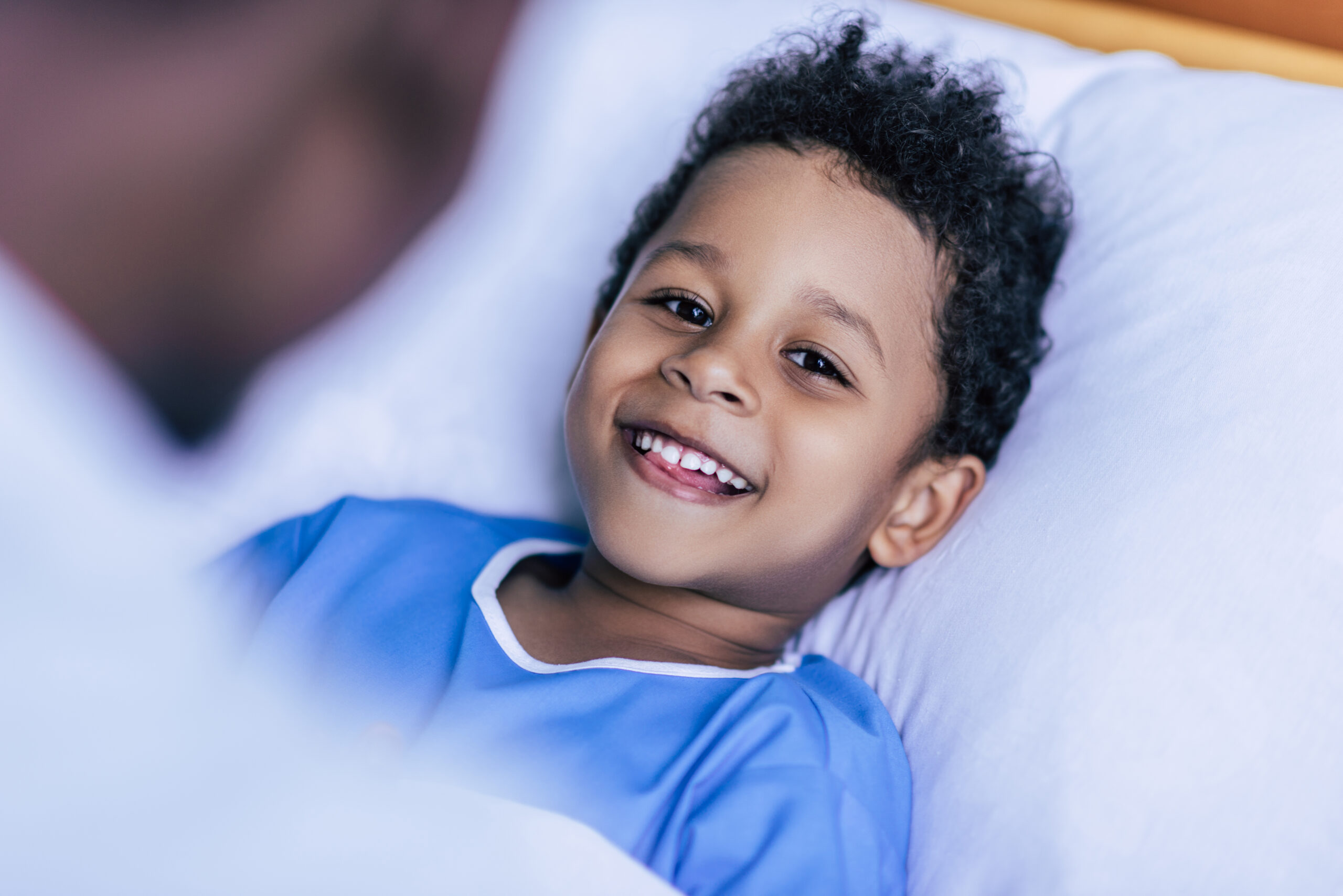 A young child wearing a blue hospital gown lies in bed, smiling warmly at someone nearby, creating a joyful and comforting atmosphere.