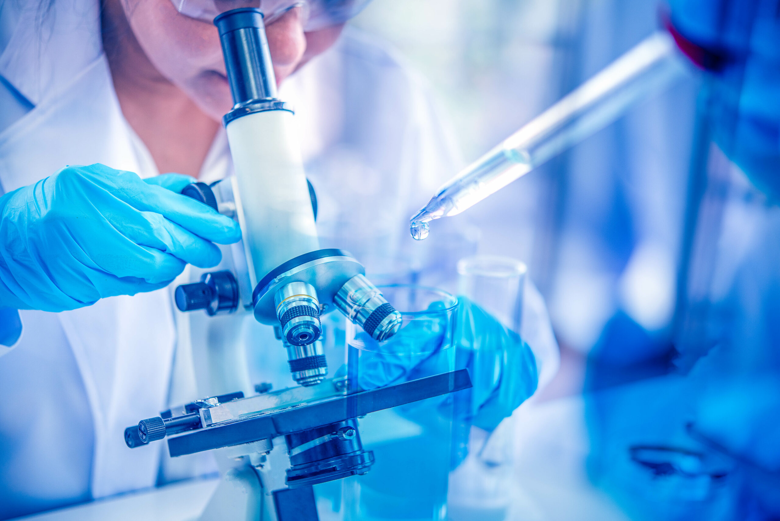 A scientist wearing blue gloves uses a microscope while another hand holds a pipette with a droplet, in a laboratory setting with glassware in the background.