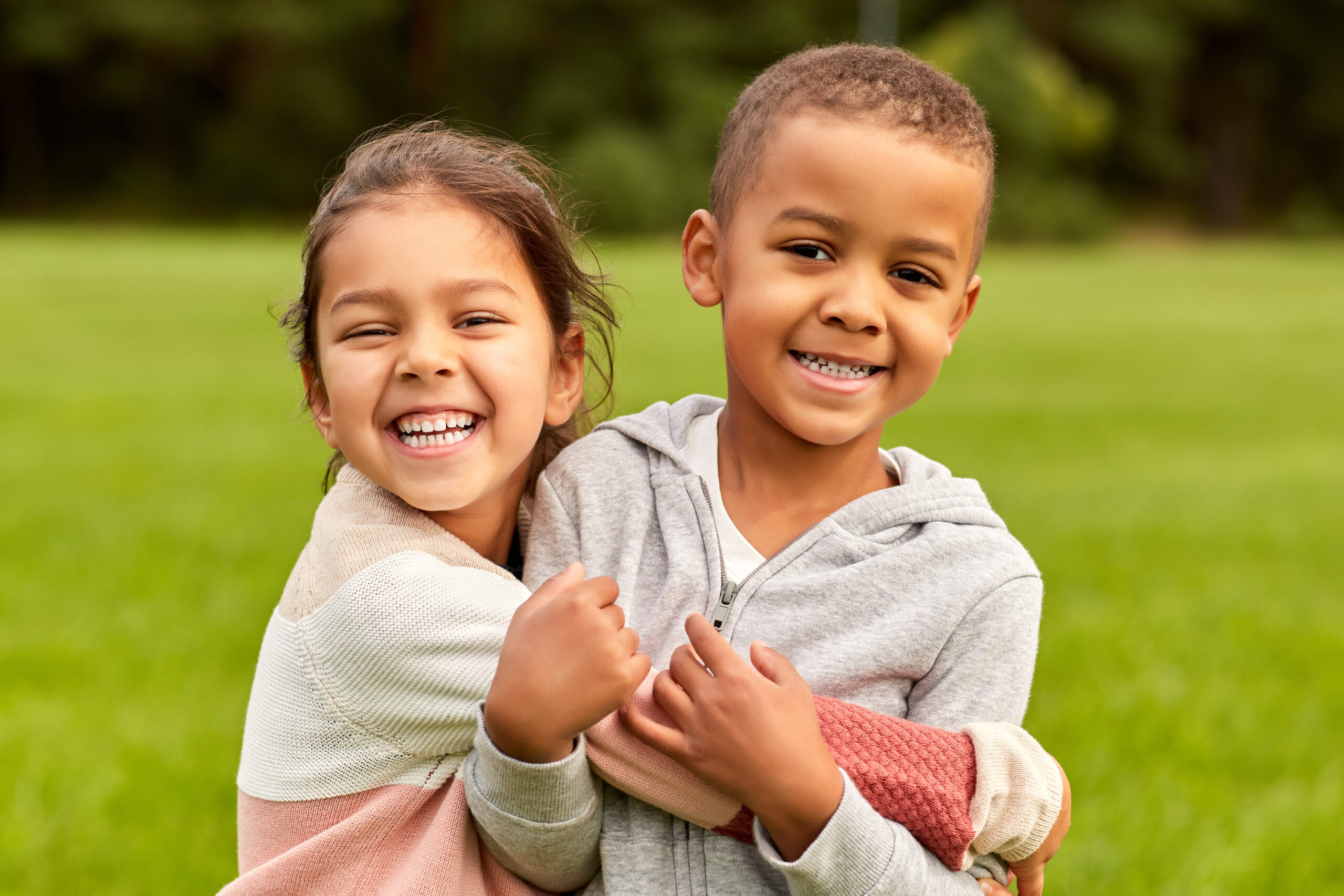 Two young children, a girl and a boy, stand close together outdoors on a grassy field, smiling and hugging each other while looking at the camera. The background is green and blurred.