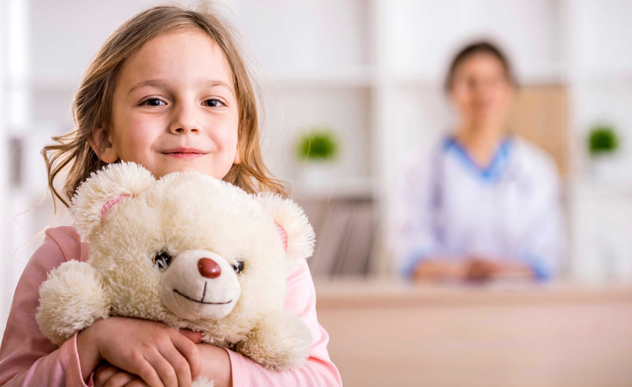 A young girl smiles and hugs a white teddy bear, standing in the foreground. In the background, a woman in a white coat, possibly a doctor or nurse, is sitting at a desk in a bright room.