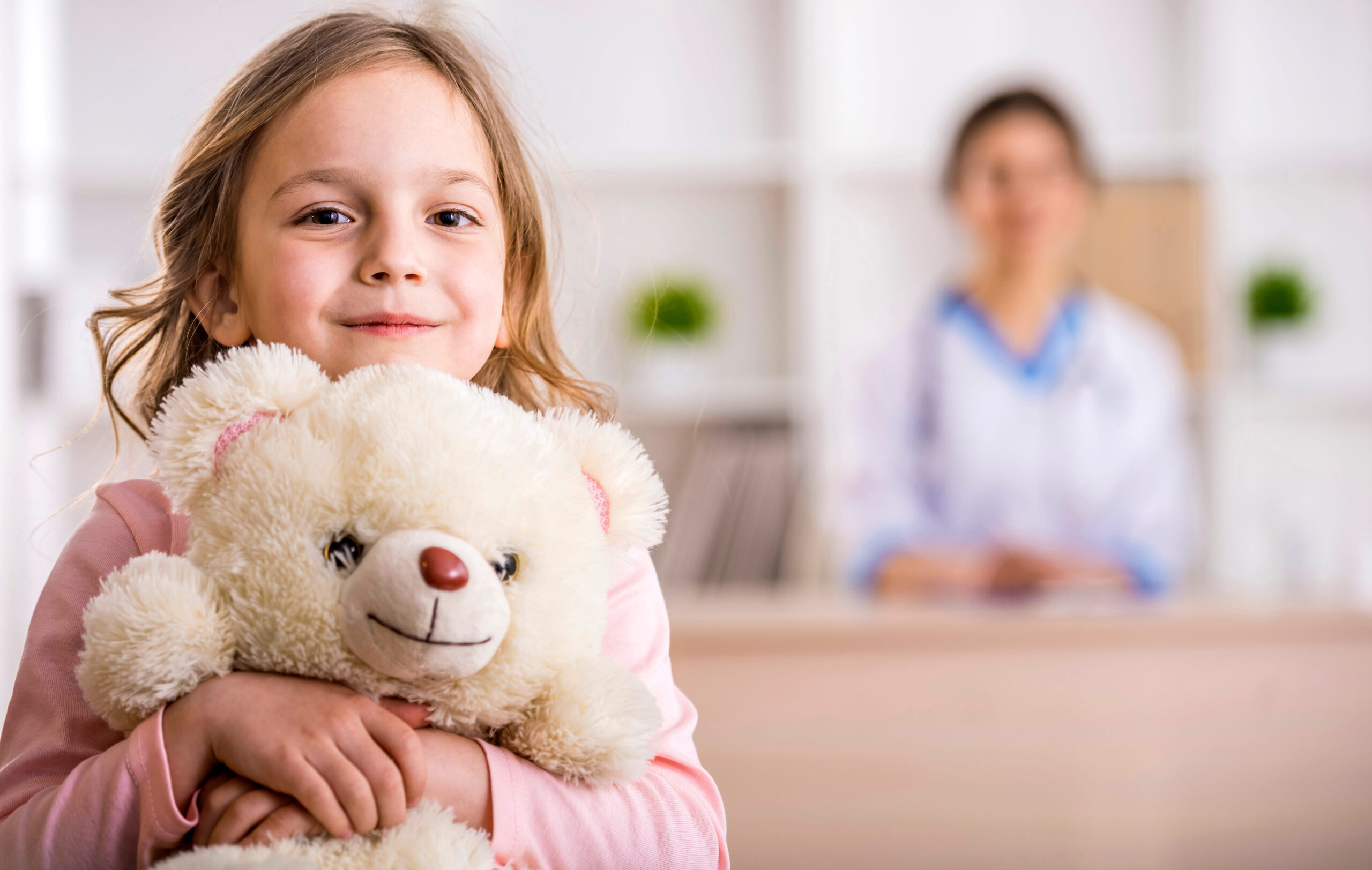 A young girl smiles and hugs a white teddy bear in the foreground, while a woman in a white coat, likely a doctor, sits at a desk in a blurred background of a bright, welcoming office.