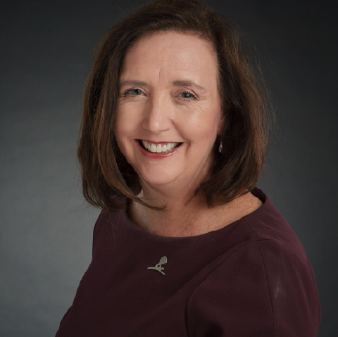 A woman with shoulder-length brown hair smiles at the camera. She is wearing a burgundy top with a small silver pin and pearl earrings, set against a dark, plain background.