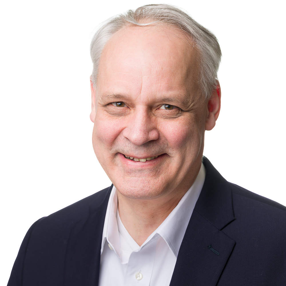 A middle-aged man with light skin, short gray hair, and wearing a dark suit jacket over a white collared shirt, smiling against a plain white background.