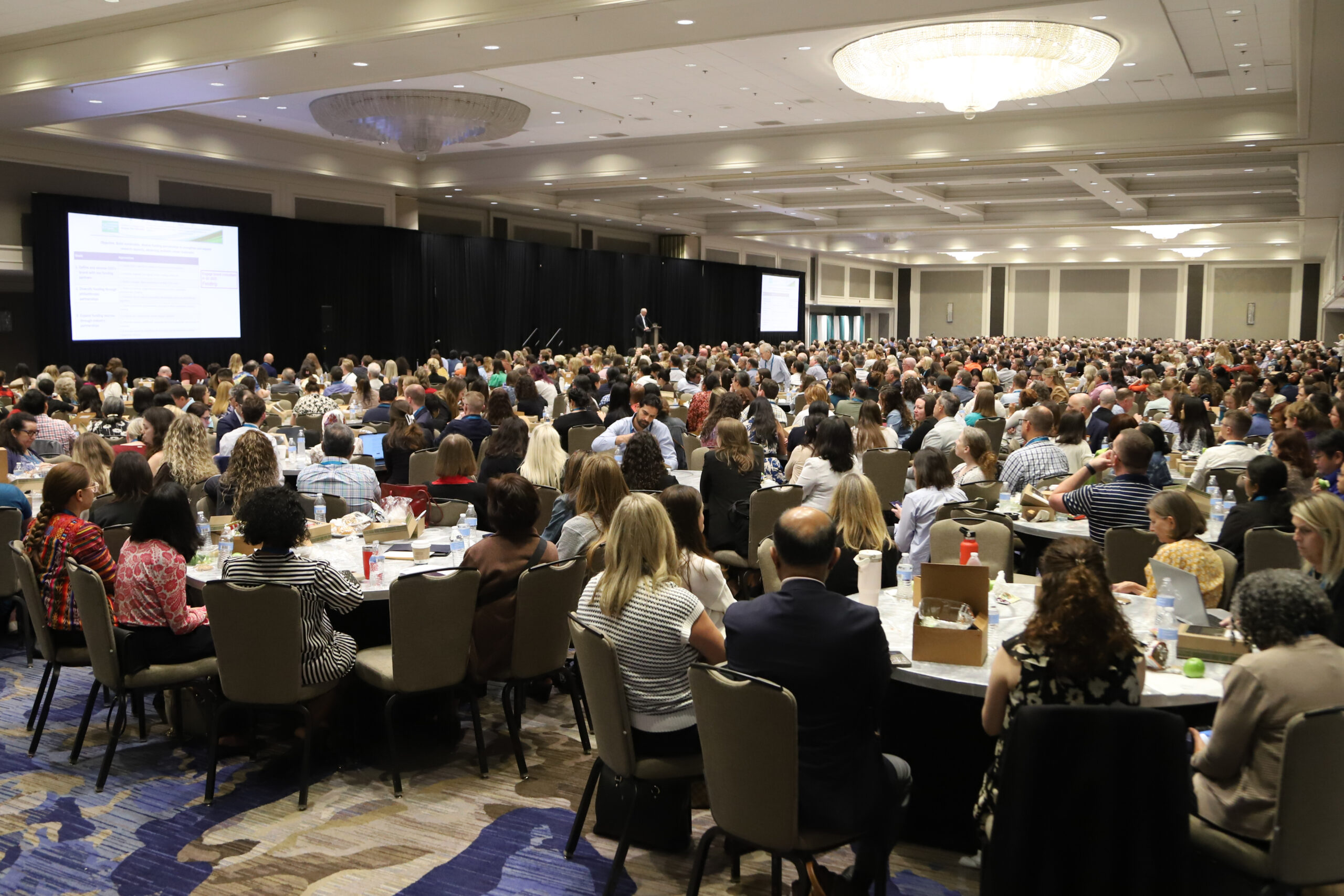 A large conference room filled with people seated at round tables, facing a stage with a speaker and presentation screens. The room is brightly lit with chandeliers and decorated in neutral colors.