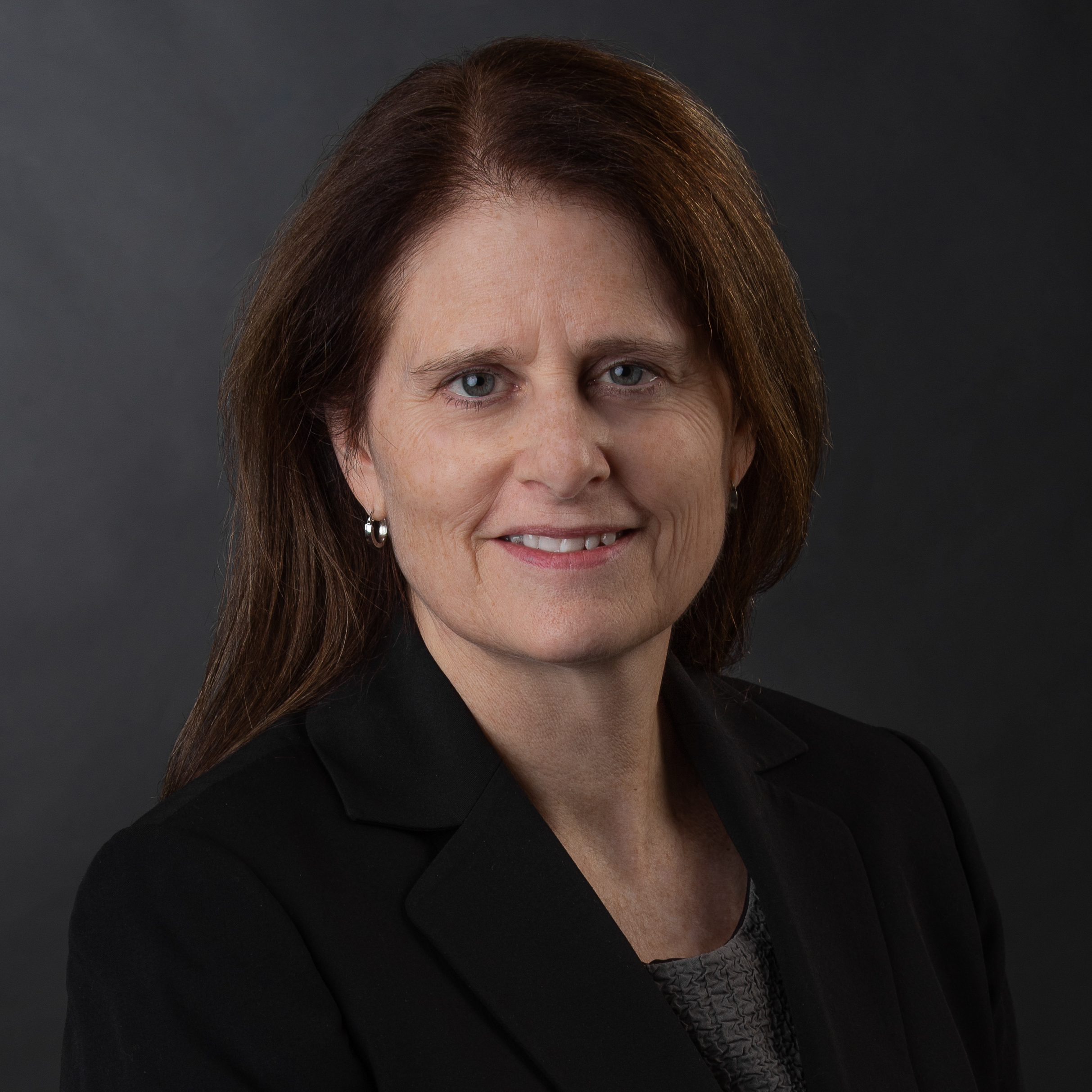 A woman with long brown hair wearing a black blazer and gray top poses in front of a dark background, smiling slightly at the camera.