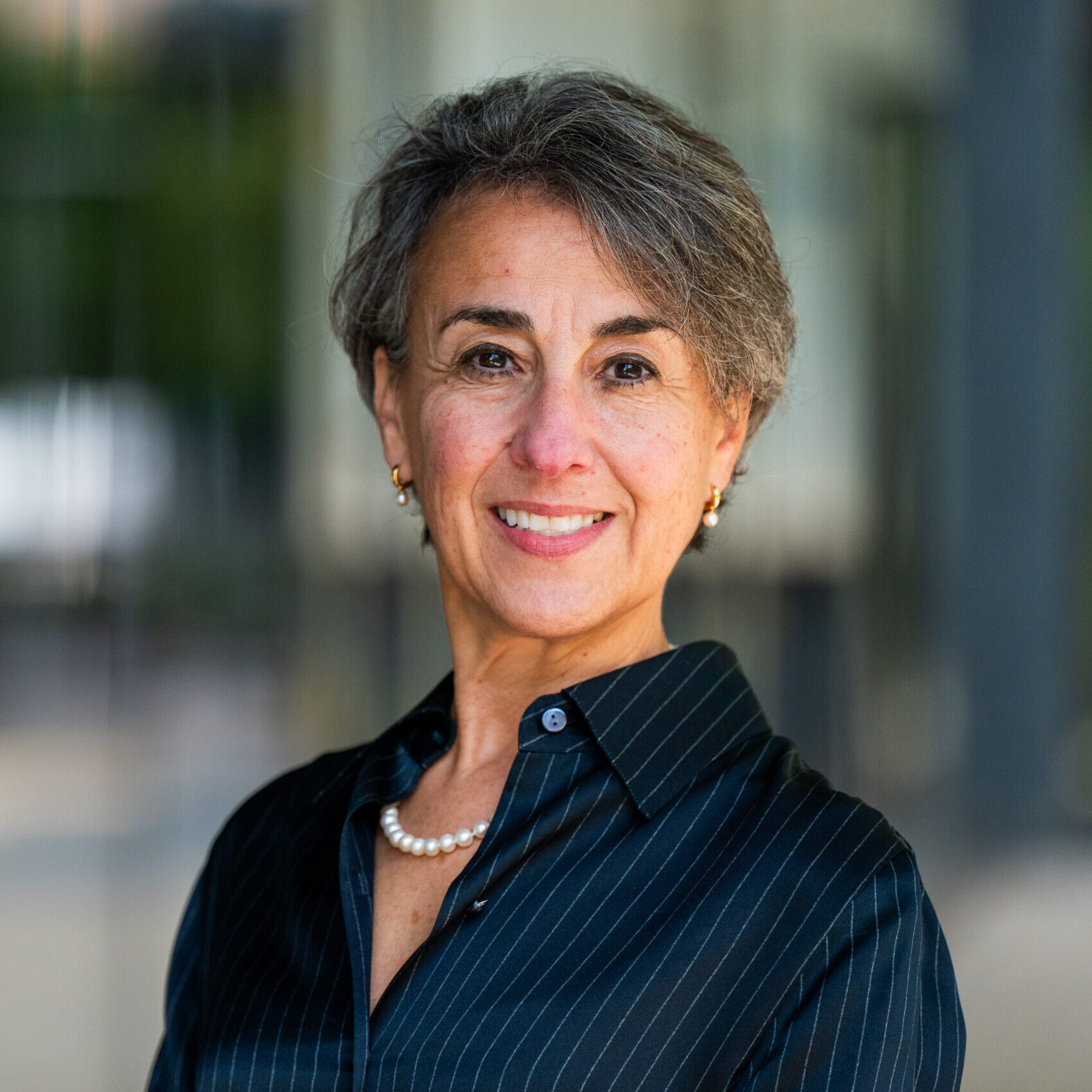 A woman with short gray hair smiles at the camera. She is wearing a dark pinstripe blouse, a pearl necklace, and small earrings, standing in front of a blurred, neutral background.