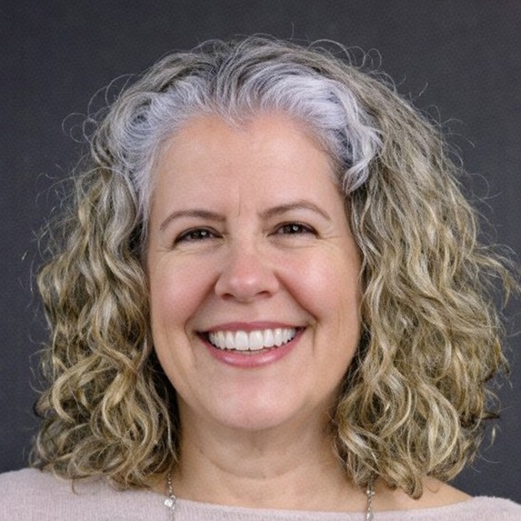 A middle-aged woman with curly, shoulder-length gray and blonde hair smiles warmly at the camera. She is wearing a light-colored top and a necklace, with a plain dark background behind her.