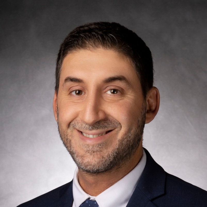 A man with short dark hair and a trimmed beard, wearing a dark suit, white shirt, and tie, smiles at the camera against a gray studio background.