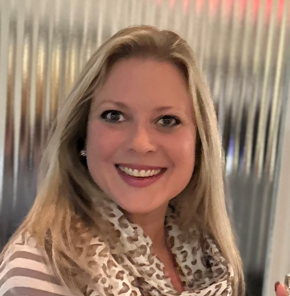 A smiling woman with long blonde hair wearing a leopard-print scarf and a striped top, standing in front of a blurred, metallic background.