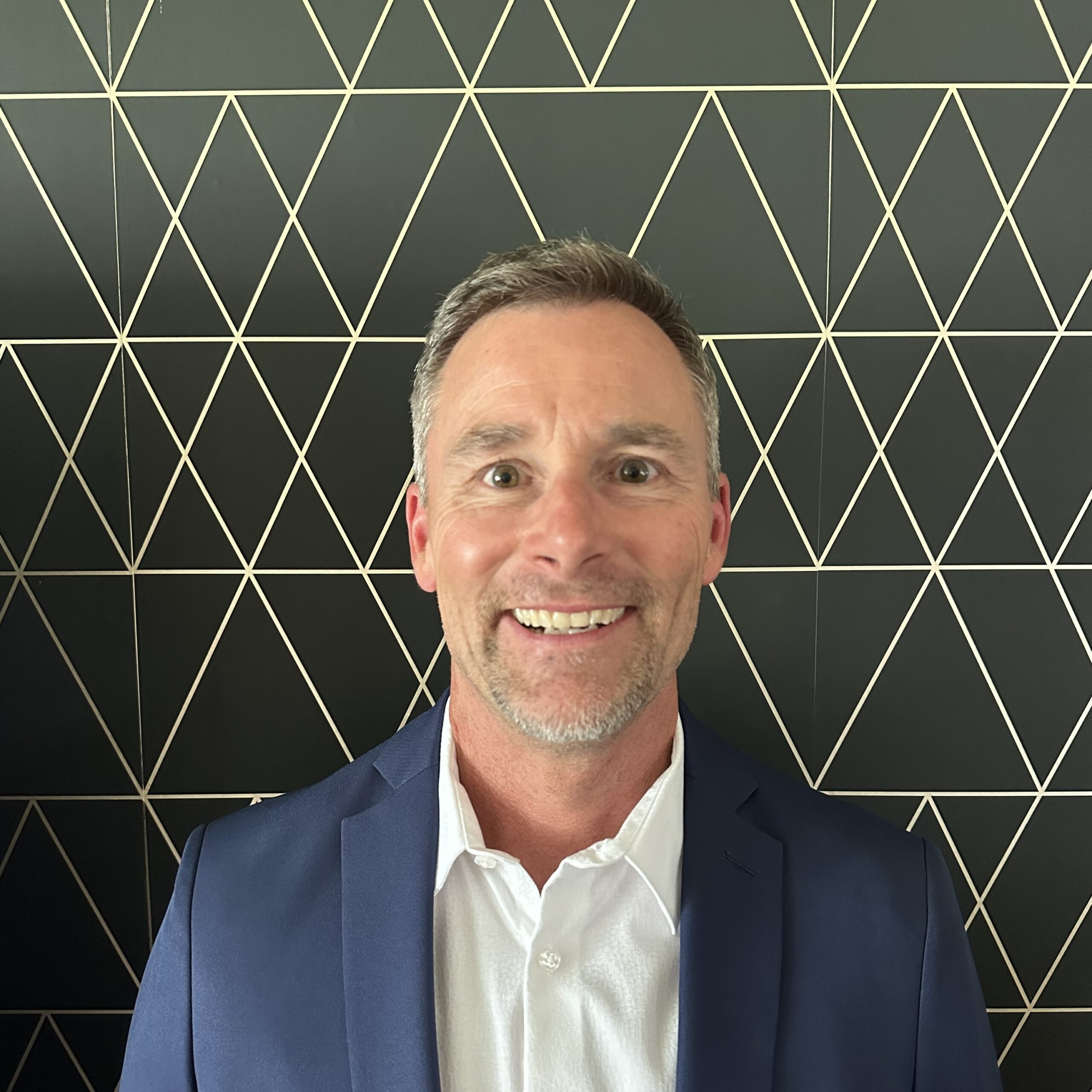 A smiling man with short gray hair and a trimmed beard wears a blue blazer over a white shirt, standing in front of a black wall with a gold geometric triangle pattern.