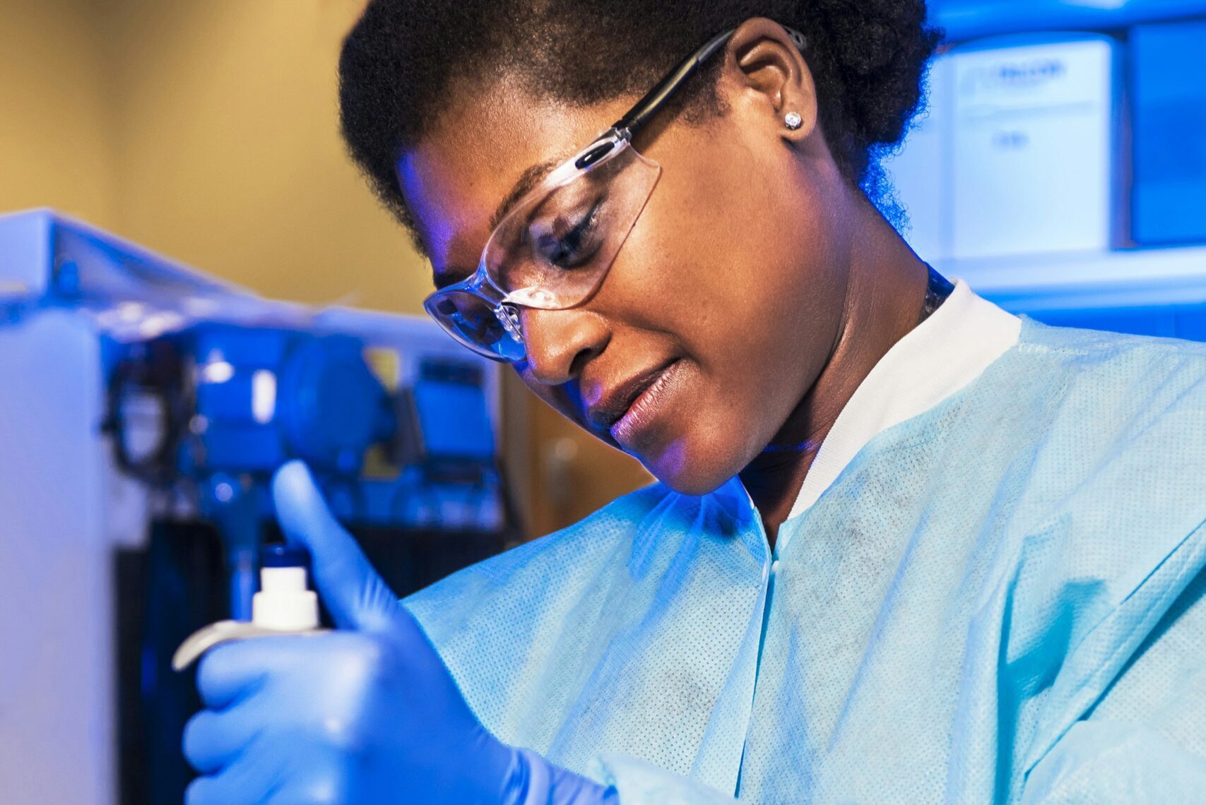 A scientist wearing safety glasses, gloves, and a lab coat carefully uses a pipette in a laboratory setting with blue lighting and scientific equipment in the background.