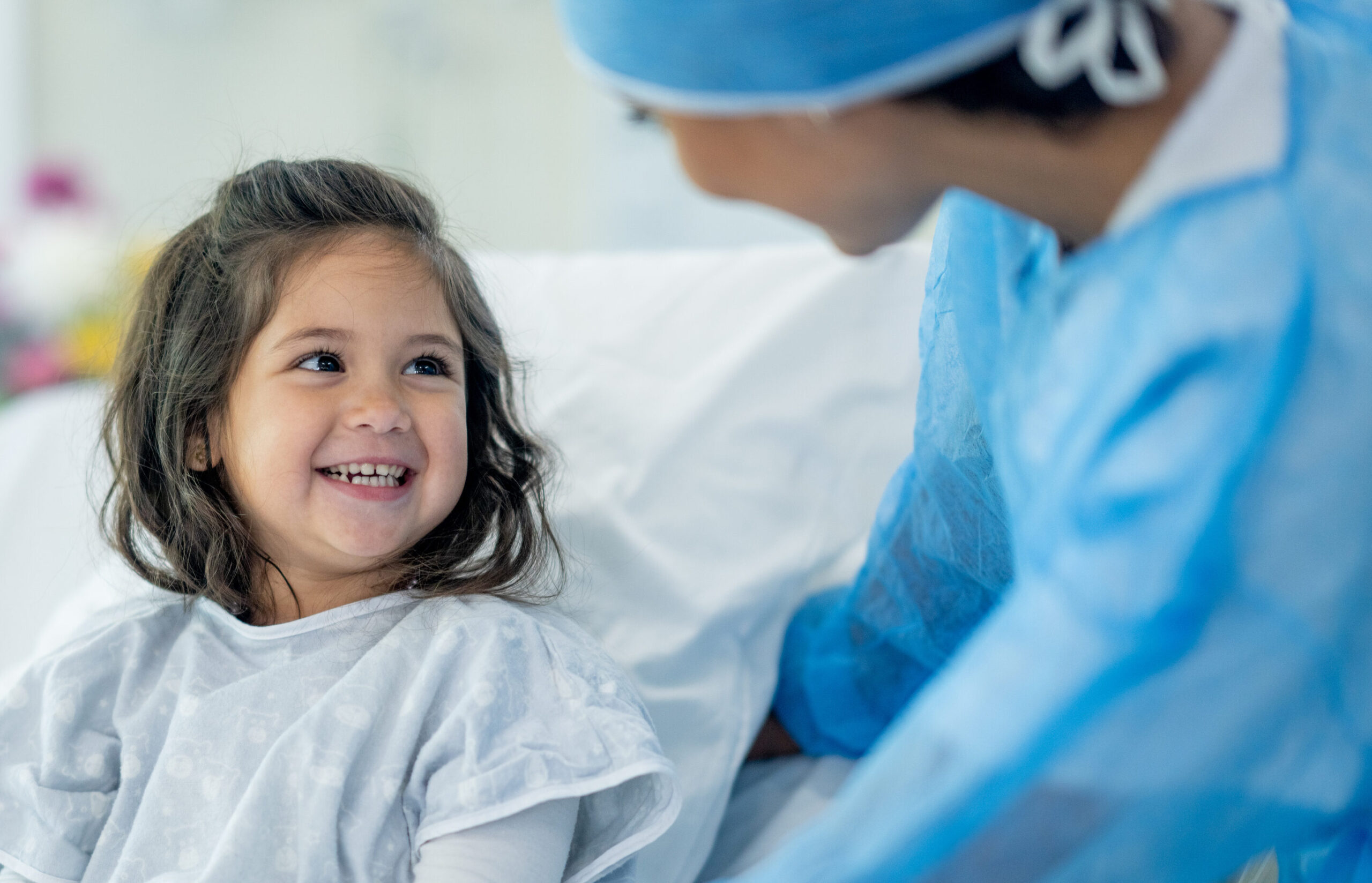 A young girl in a hospital gown smiles happily while sitting in a hospital bed, looking up at a healthcare worker in blue scrubs and a cap, who is leaning towards her.