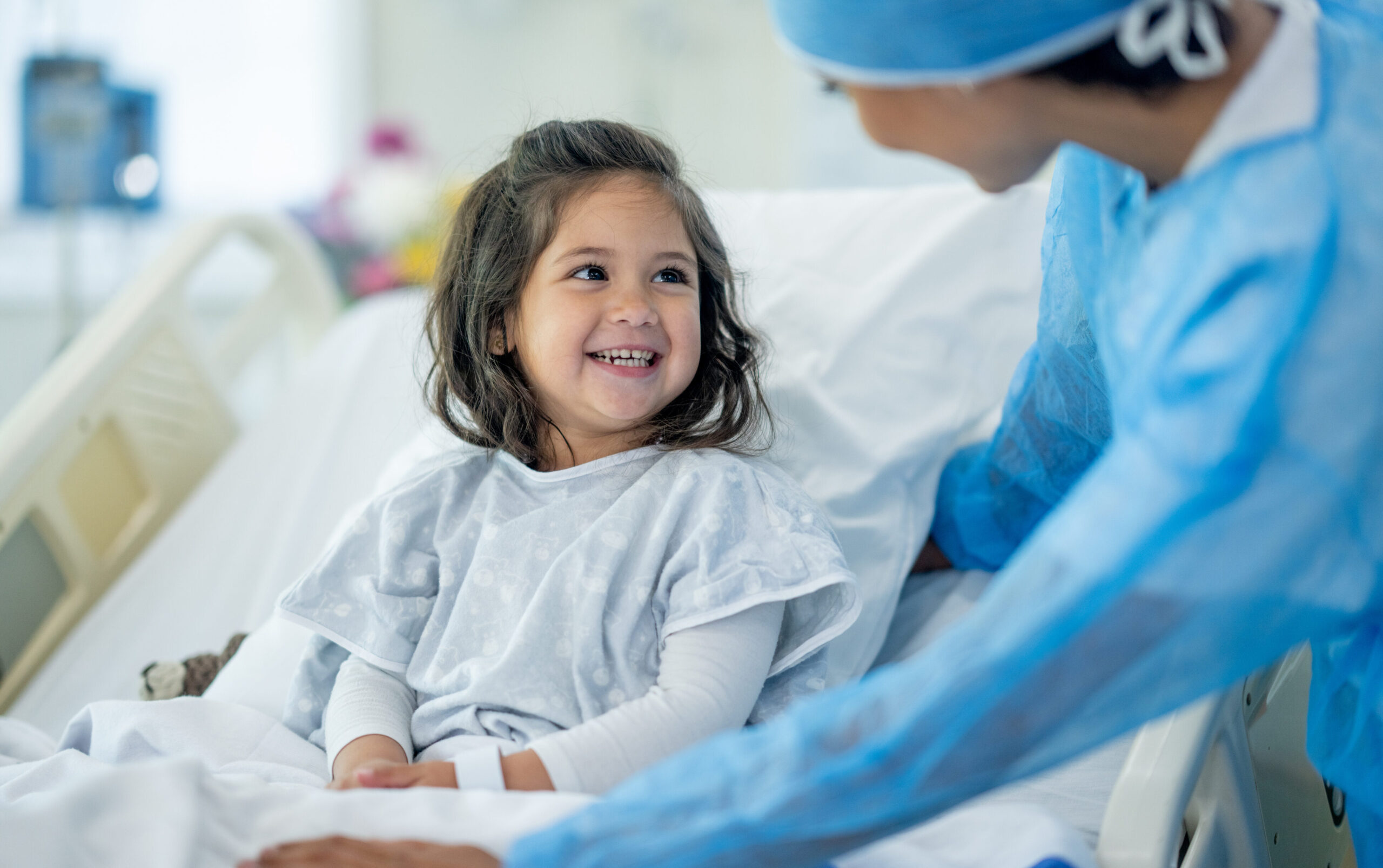 A young child smiles while sitting in a hospital bed, wearing a hospital gown. A healthcare worker in blue scrubs and a mask leans toward the child, offering comfort and support.