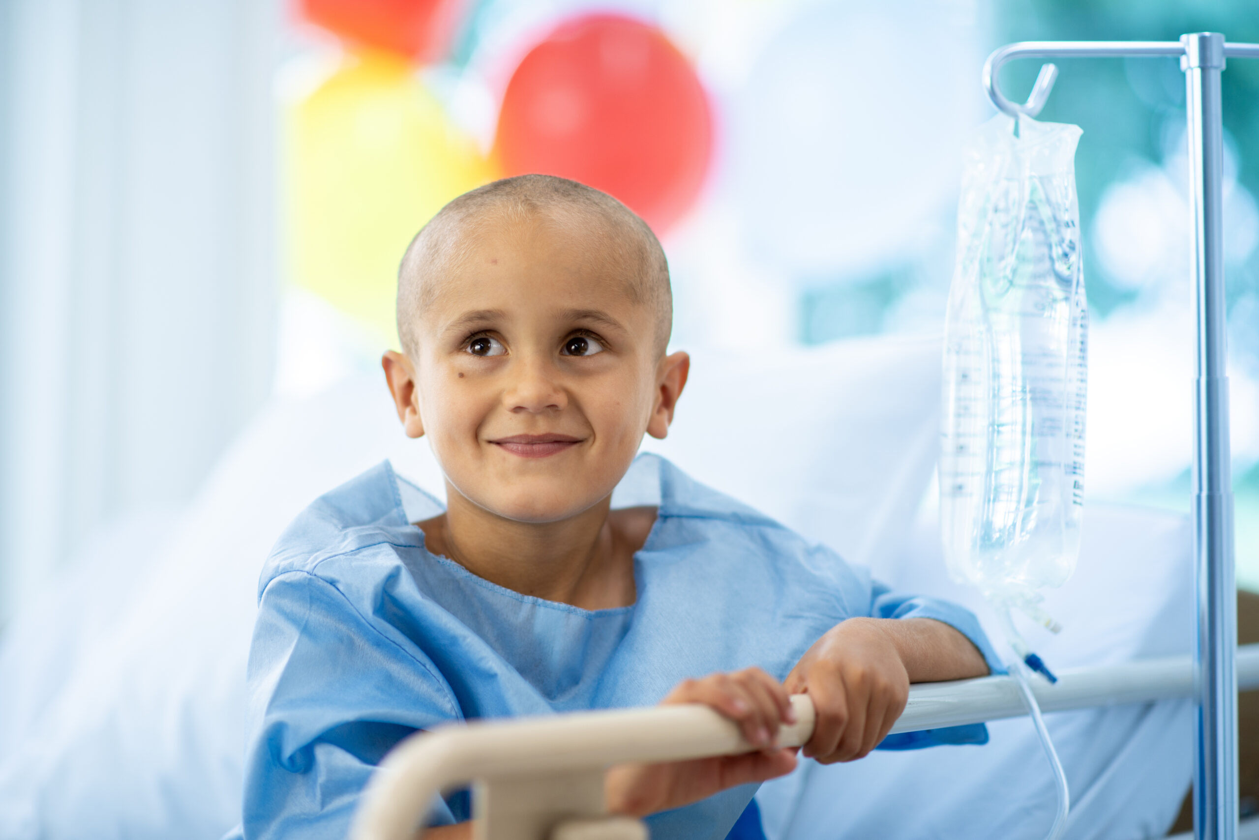 A young child with a bald head, wearing a hospital gown, smiles while sitting in a hospital bed. An IV drip is nearby, and colorful balloons are visible in the background.
