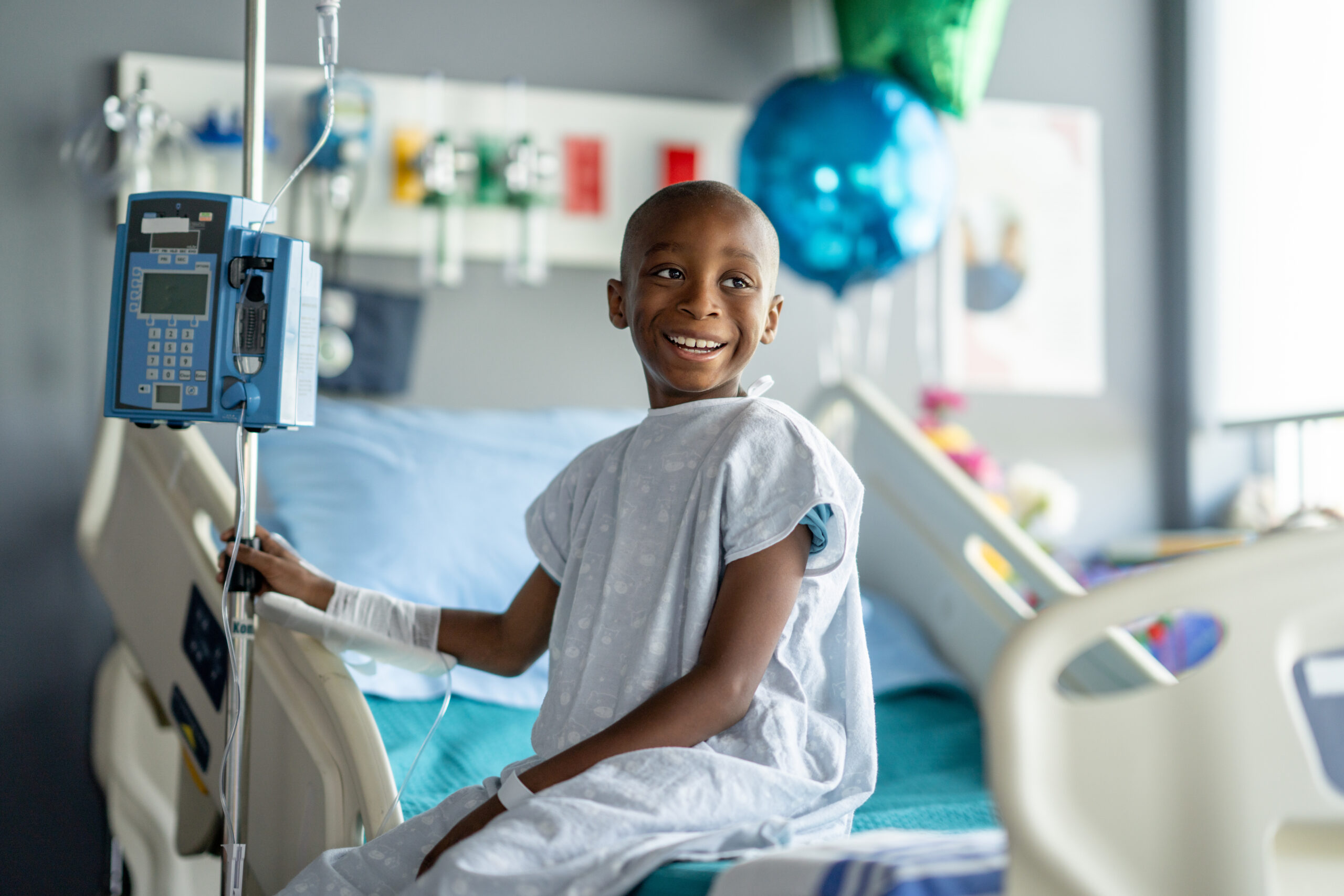 A smiling child wearing a hospital gown sits on a hospital bed, holding onto an IV pole. Medical equipment and blue balloons are visible in the background, creating a bright and hopeful atmosphere.
