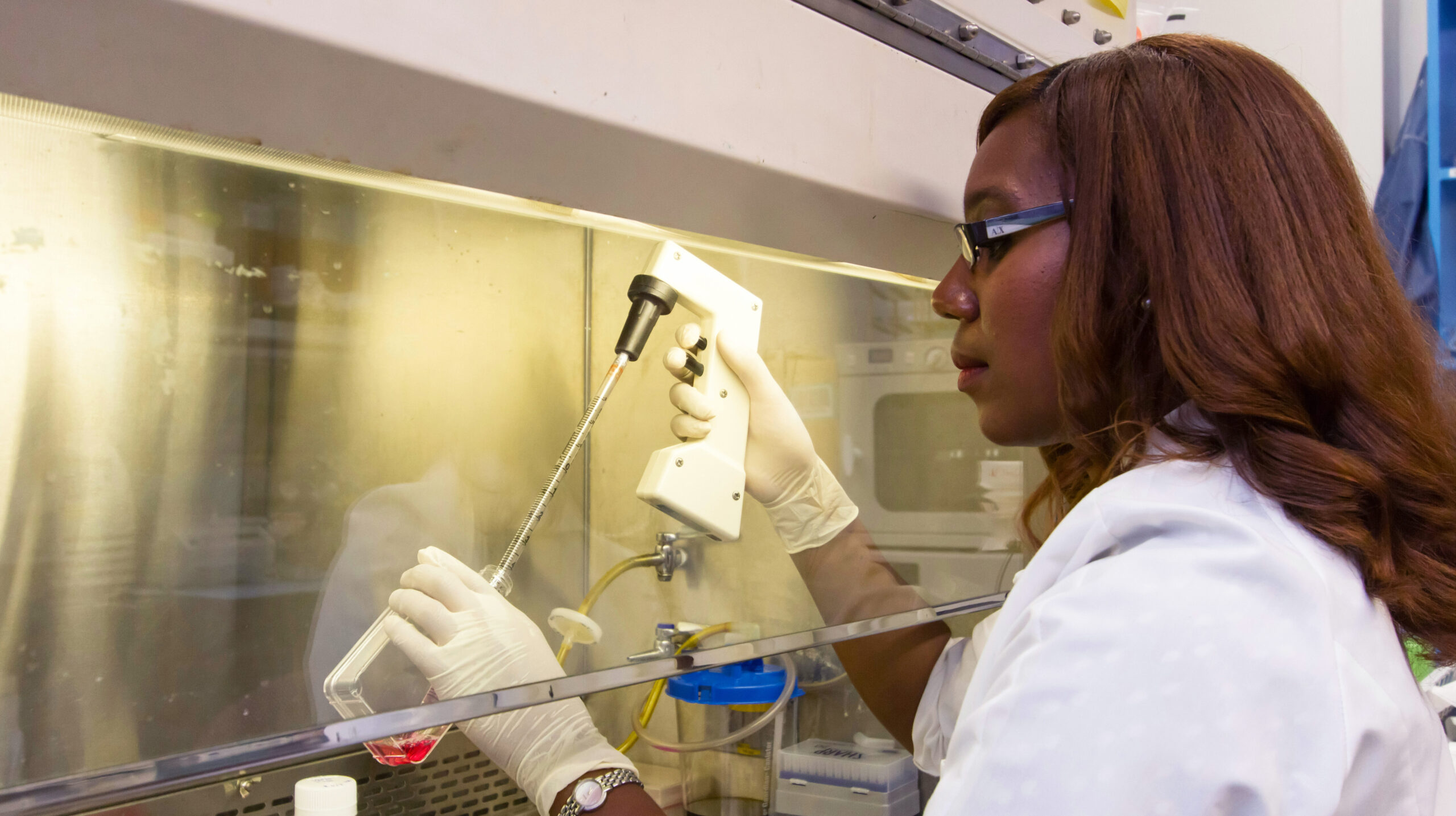 A scientist wearing a lab coat, gloves, and safety glasses uses a pipette to transfer liquid into a container inside a laboratory fume hood.