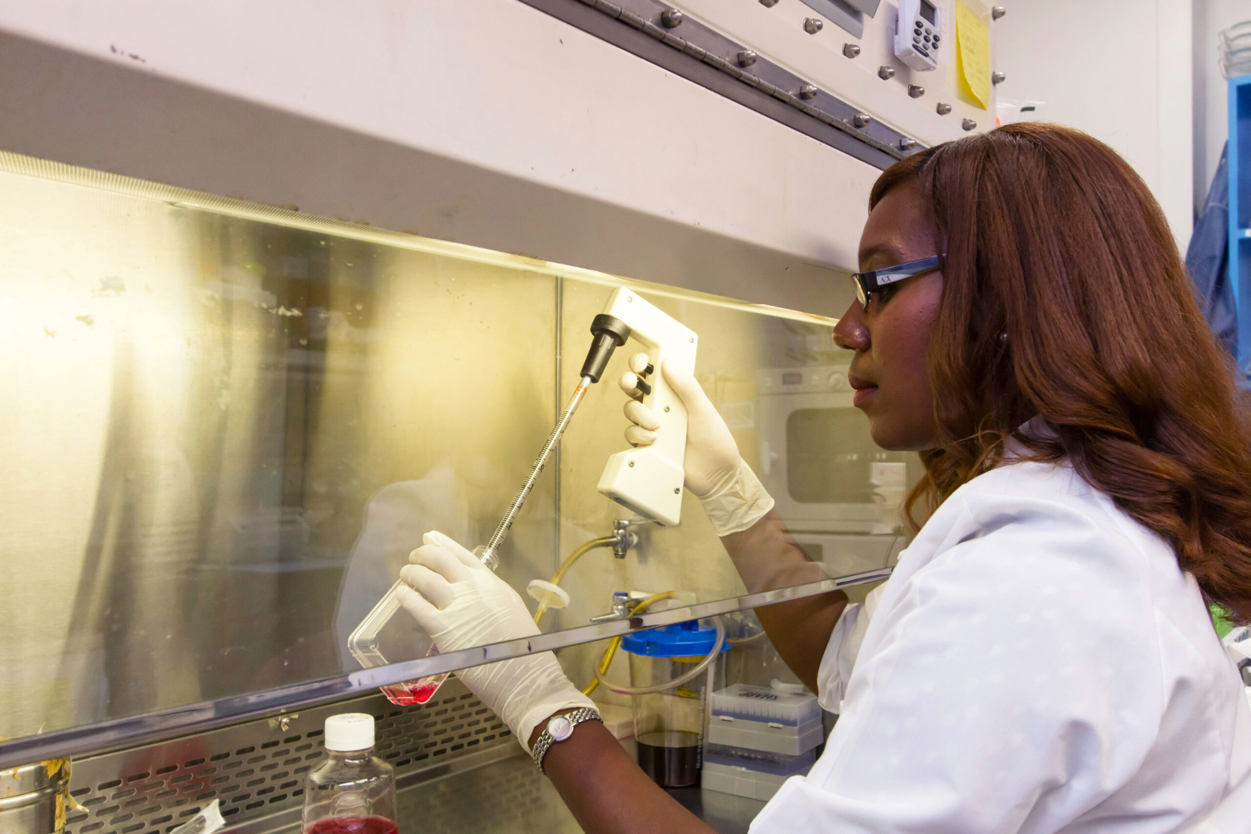 A scientist wearing a white lab coat and gloves uses a pipette to transfer liquid into a container inside a laboratory fume hood. Various lab equipment and bottles are visible on the work surface.