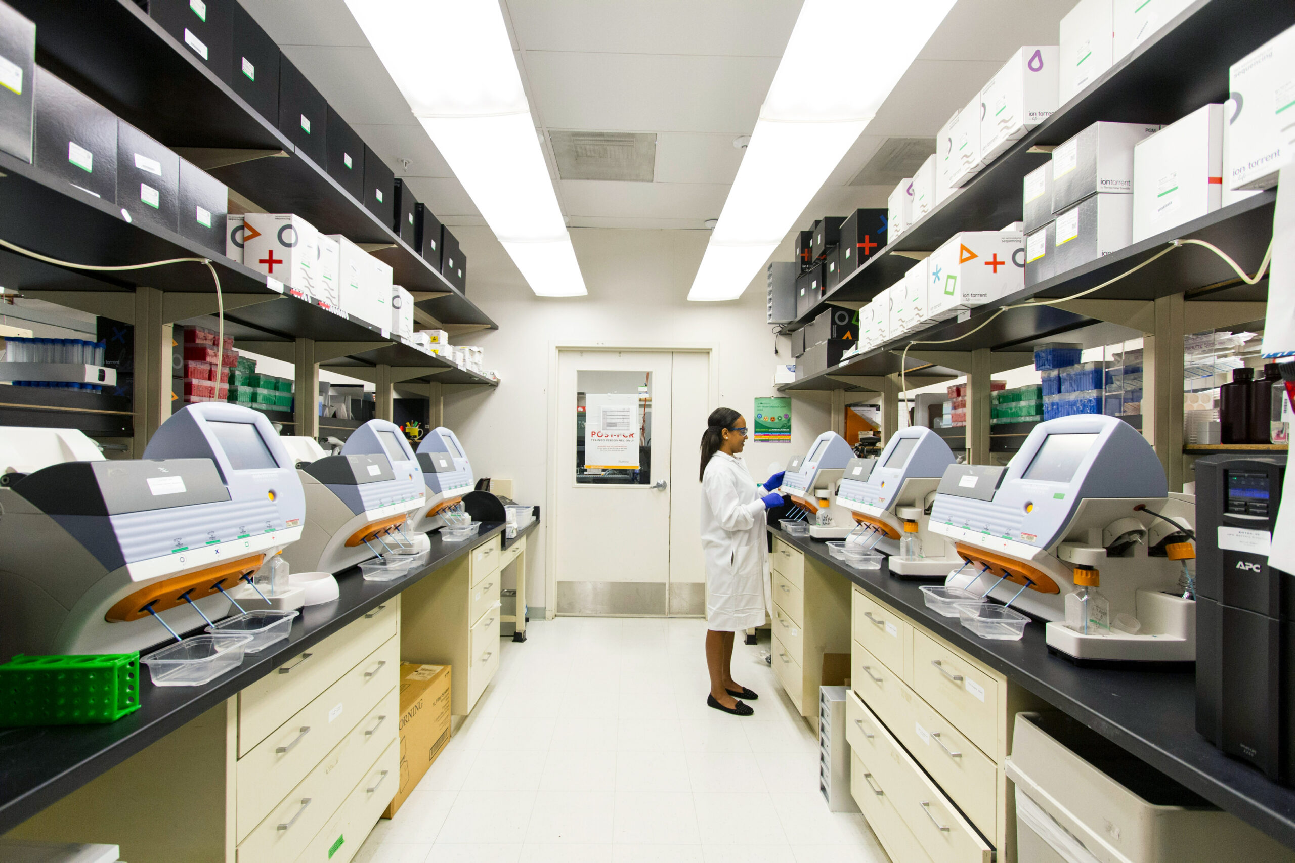 A person in a white lab coat stands in a bright laboratory filled with equipment, machines, and organized shelves holding boxes and supplies. The person appears to be working with a machine.