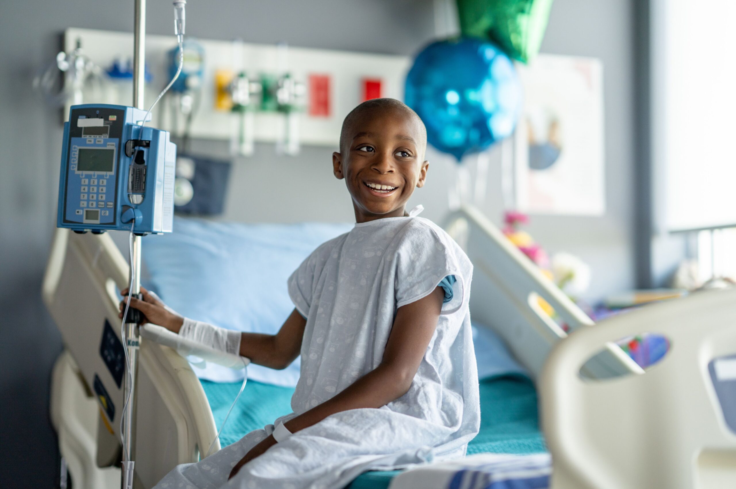 Smiling child receiving cancer treatment while sitting beside an IV infusion pump in a hospital room.
